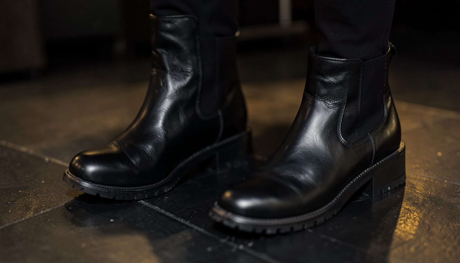 A close-up of black leather ankle boots and chunky Chelsea boots on a dark floor with subtle golden highlights