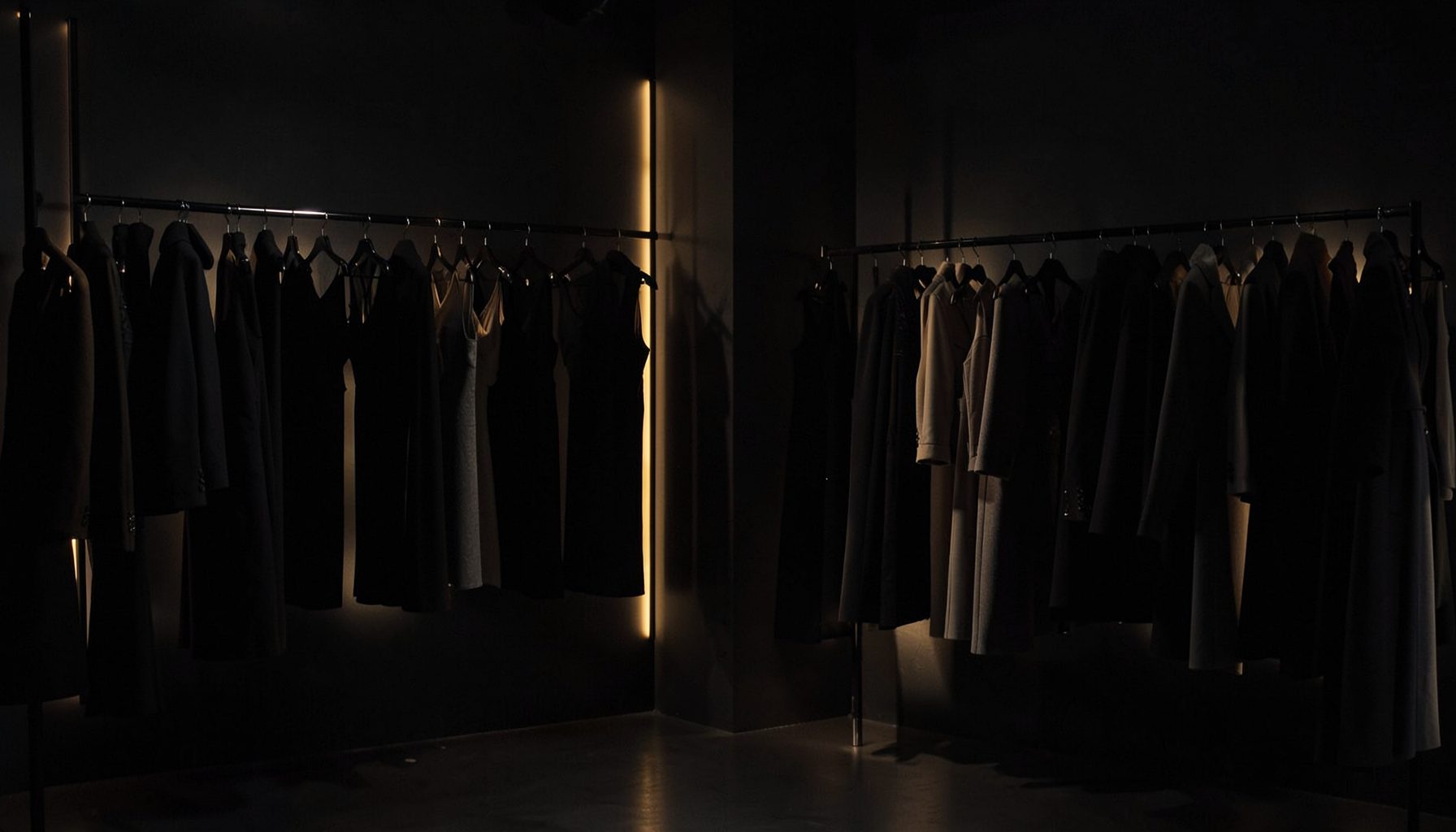 A moody fashion editorial scene with dark charcoal and black tones, soft golden accent lighting, and racks of women's clothing