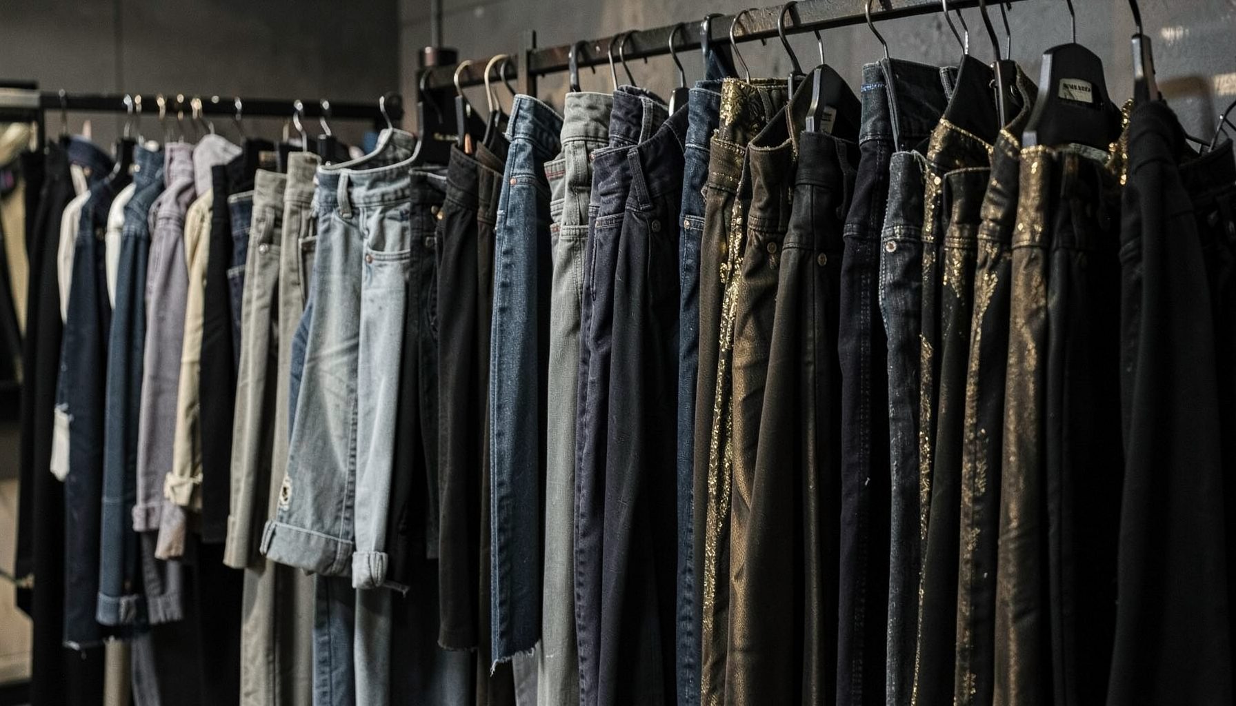 Rows of folded jeans and hanging dresses in muted grey, black and gold tones, giving a moody fashion-boutique feel